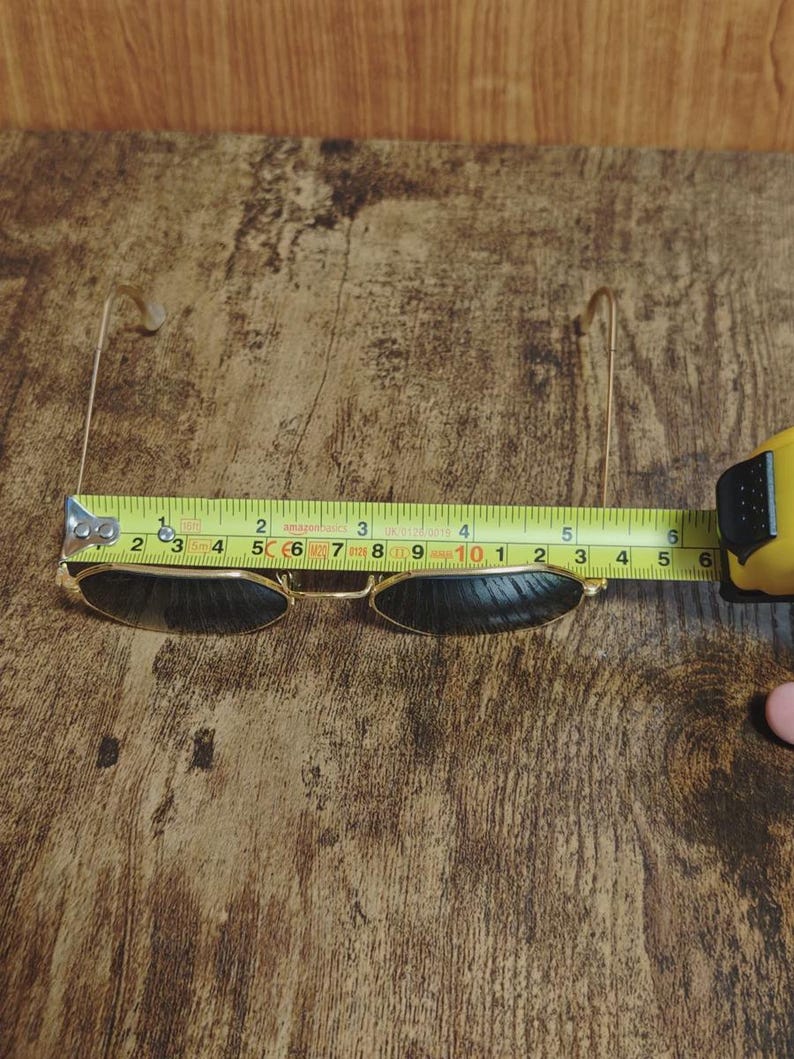 May include: Gold-framed sunglasses with dark lenses are measured with a yellow tape measure. The sunglasses are approximately 5 inches wide. The background is a wooden surface.