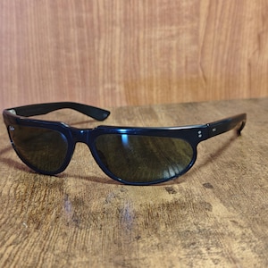 May include: Black sunglasses with a curved frame and dark lenses rest on a wooden surface. The sunglasses have a sleek, modern design, with a thick frame and a subtle curve at the top. The lenses appear to be tinted, providing shade and protection from the sun.
