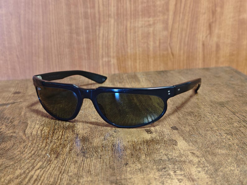 May include: Black sunglasses with a curved frame and dark lenses rest on a wooden surface. The sunglasses have a sleek, modern design, with a thick frame and a subtle curve at the top. The lenses appear to be tinted, providing shade and protection from the sun.