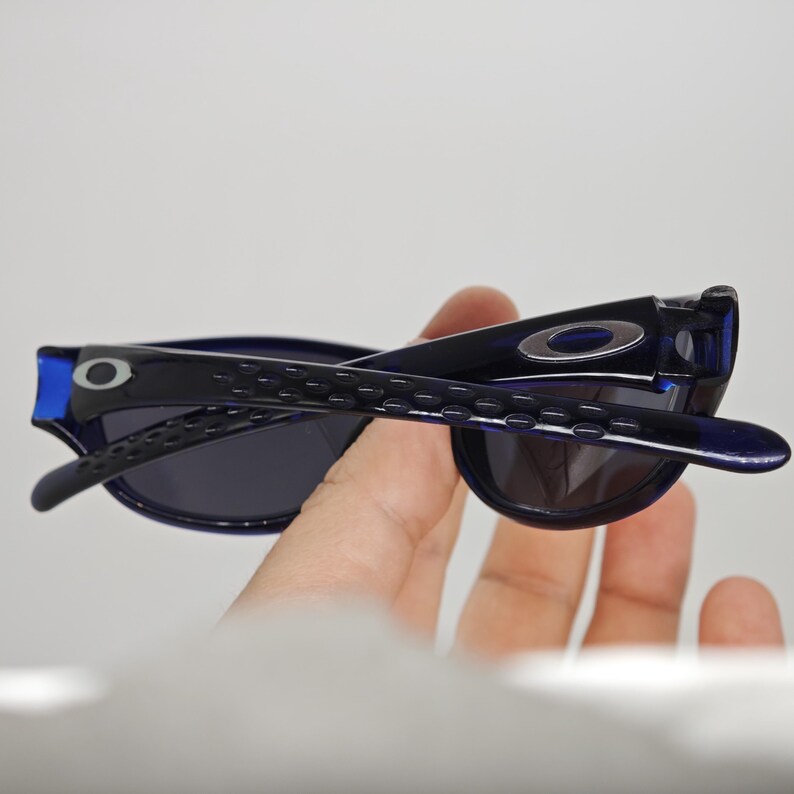 May include: Blue and black sunglasses with dark lenses. The sunglasses have a sleek design with the brand logo visible on the temples. The frame is a combination of blue and black plastic.