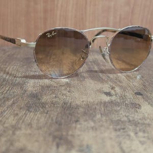 Peut inclure: Lunettes de soleil Ray-Ban dor&eacute;es avec verres d&eacute;grad&eacute;s marron. Les montures rondes ont un double pont et des branches &agrave; motifs. Le logo Ray-Ban est visible sur le verre sup&eacute;rieur gauche.