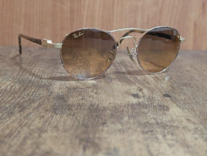Peut inclure: Lunettes de soleil Ray-Ban dor&eacute;es avec verres d&eacute;grad&eacute;s marron. Les montures rondes ont un double pont et des branches &agrave; motifs. Le logo Ray-Ban est visible sur le verre sup&eacute;rieur gauche.