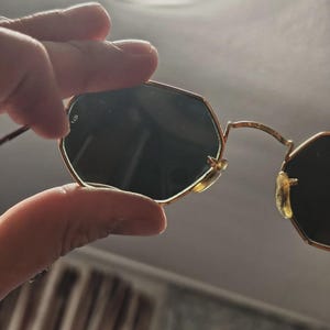May include: Gold-framed sunglasses with dark lenses. The sunglasses have a unique octagonal shape. The frame is thin and delicate, with a classic design. The lenses are dark, providing shade and protection from the sun.