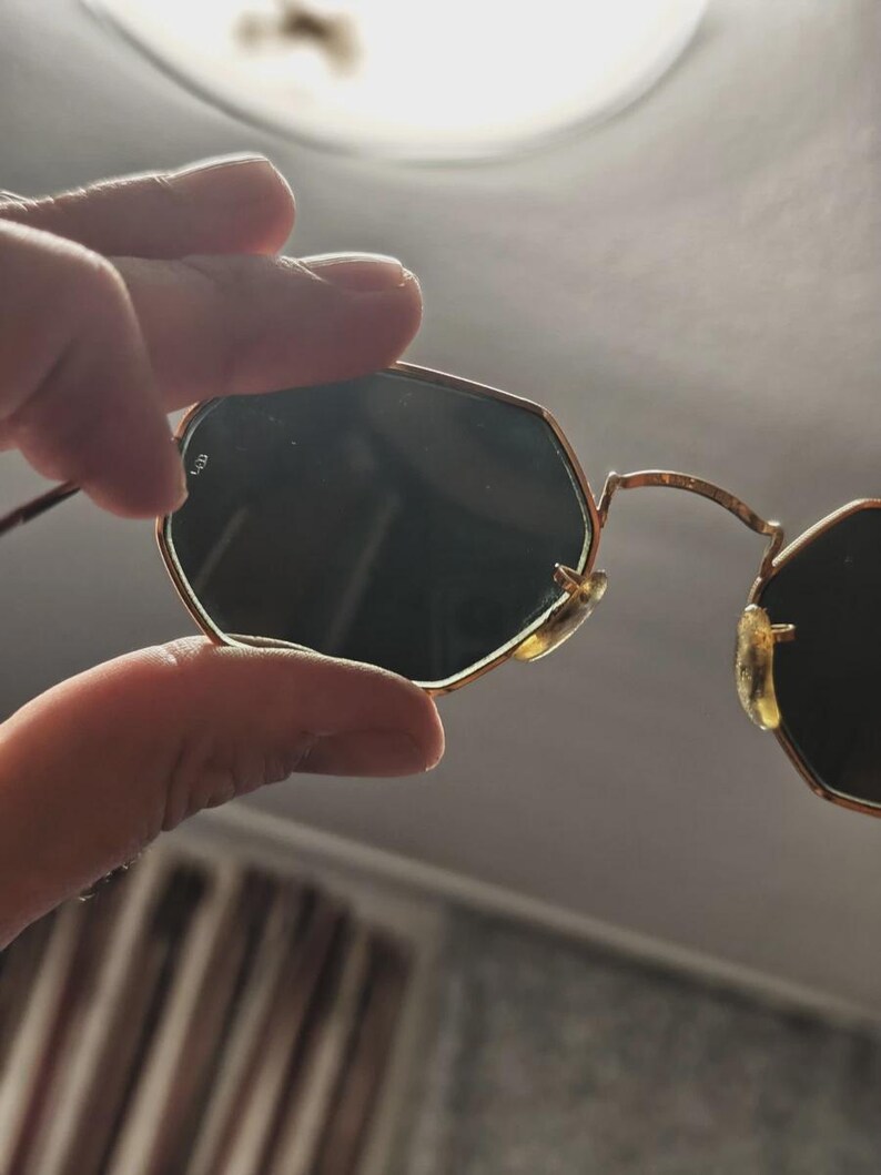 May include: Gold-framed sunglasses with dark lenses. The sunglasses have a unique octagonal shape. The frame is thin and delicate, with a classic design. The lenses are dark, providing shade and protection from the sun.