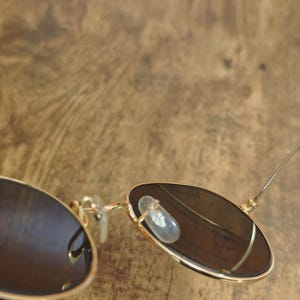 May include: Gold-framed sunglasses with dark lenses. The sunglasses have a classic aviator style with a double bridge and clear nose pads. The lenses are a dark brown color, and the frame is a shiny gold.