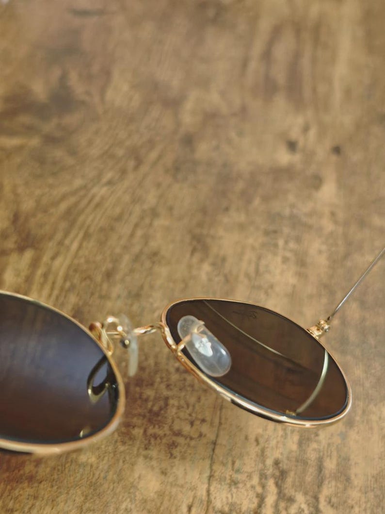 May include: Gold-framed sunglasses with dark lenses. The sunglasses have a classic aviator style with a double bridge and clear nose pads. The lenses are a dark brown color, and the frame is a shiny gold.