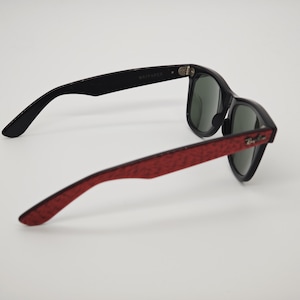 May include: Black Wayfarer sunglasses with red patterned arms. The sunglasses have dark lenses and the Ray-Ban logo is visible on the right arm. The text "WAYFARER" is printed on the left arm.