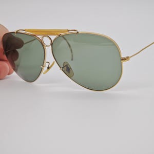 May include: Gold-framed aviator sunglasses with green lenses. The sunglasses have a double bridge and a light-colored bar across the top. The lenses are teardrop-shaped, and the arms are thin and gold-colored.