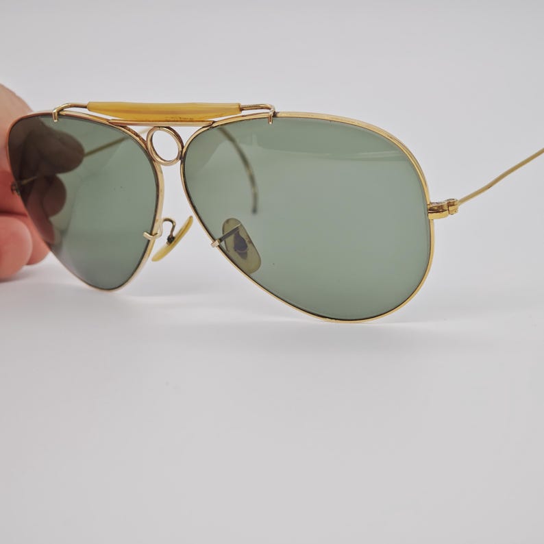 May include: Gold-framed aviator sunglasses with green lenses. The sunglasses have a double bridge and a light-colored bar across the top. The lenses are teardrop-shaped, and the arms are thin and gold-colored.