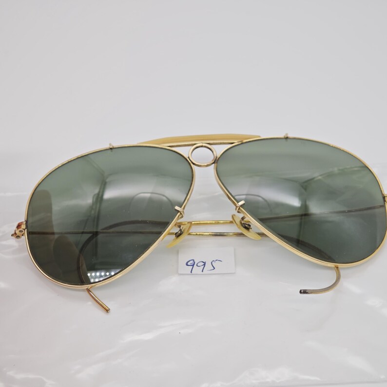 May include: Gold-framed aviator sunglasses with green lenses. The sunglasses have a double bridge and a curved brow bar. The lenses are a teardrop shape. The number "995" is on a white sticker.