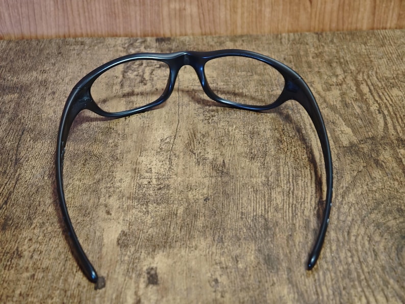 Op de afbeelding: Zwarte bril met een gebogen montuur en ovale glazen. De bril staat op een houten achtergrond. Het montuur is effen donker van kleur en de glazen zijn helder. De pootjes van de bril zijn gebogen.