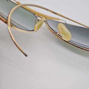 May include: Close-up of gold-rimmed sunglasses with gray lenses. The sunglasses have a classic aviator design, featuring a thin gold frame and light-colored nose pads. The temples are also gold, with a textured design.