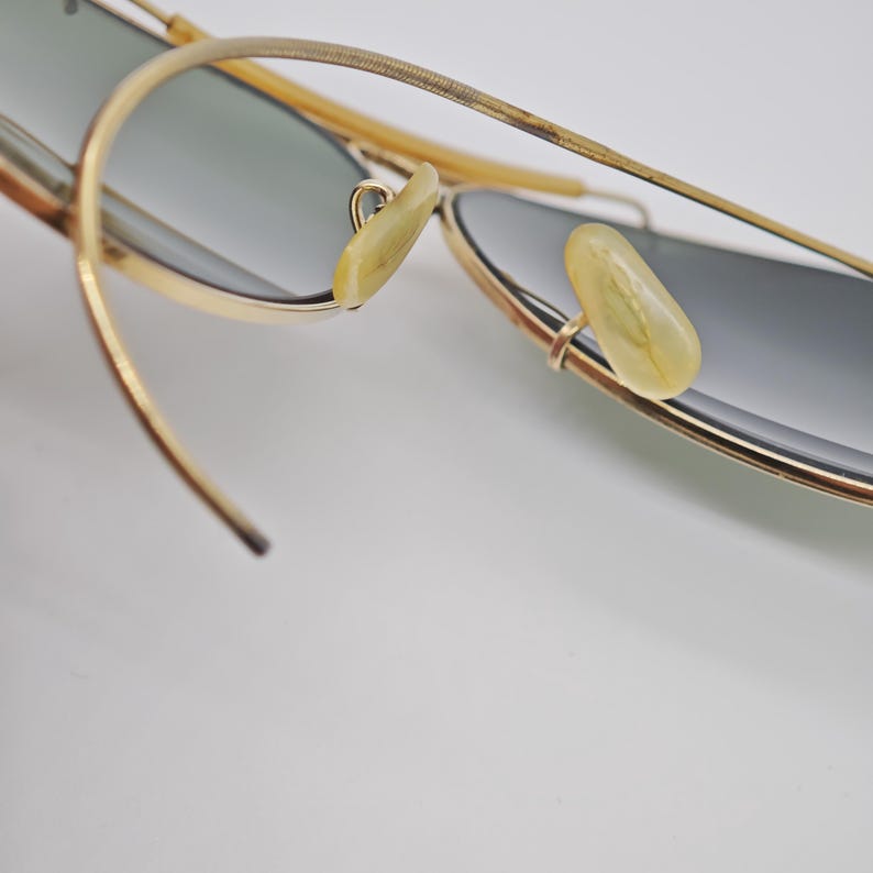 May include: Close-up of gold-rimmed sunglasses with gray lenses. The sunglasses have a classic aviator design, featuring a thin gold frame and light-colored nose pads. The temples are also gold, with a textured design.
