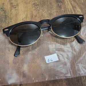 May include: Black sunglasses with a gold rim and dark lenses. The sunglasses have a classic design with a rounded shape. The arms are black and the lenses are dark. A small white tag with the number 66 is visible.