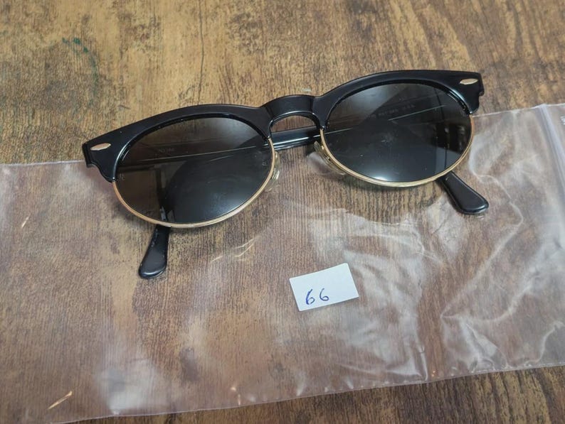 May include: Black sunglasses with a gold rim and dark lenses. The sunglasses have a classic design with a rounded shape. The arms are black and the lenses are dark. A small white tag with the number 66 is visible.