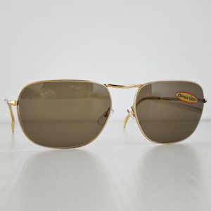 May include: Gold-tone metal frame sunglasses with brown tinted lenses. The right lens features an oval sticker with the text 'Orma 1000' in red and yellow. The sunglasses have a classic bridge design and thin metal temples with curved tips.