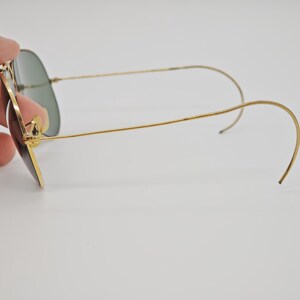 May include: Gold-framed sunglasses with green lenses. The glasses have a classic aviator shape, with a thin gold frame and curved temples. The lenses are a dark green color, and the overall design is sleek and stylish.