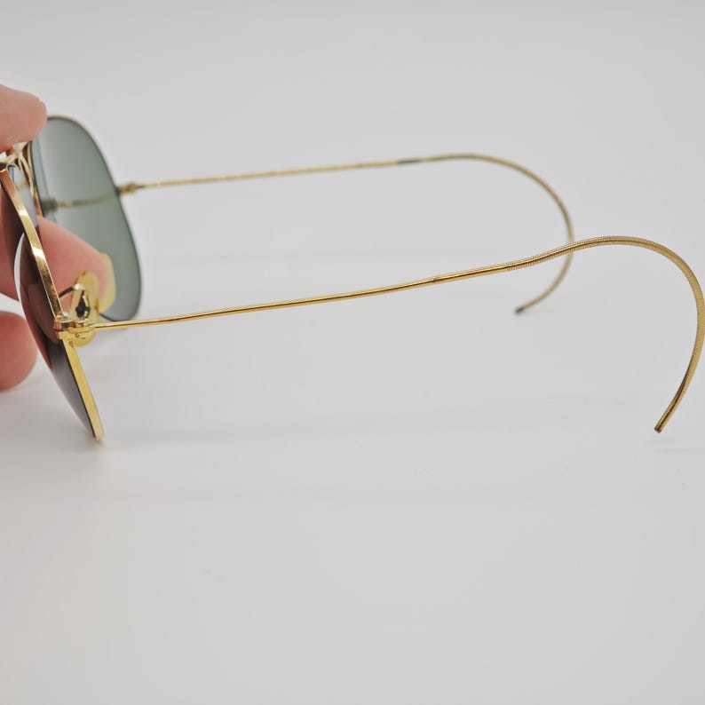 May include: Gold-framed sunglasses with green lenses. The glasses have a classic aviator shape, with a thin gold frame and curved temples. The lenses are a dark green color, and the overall design is sleek and stylish.