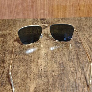 Puede incluir: Gafas de sol con montura dorada y lentes oscuras. Las gafas tienen un dise&ntilde;o cl&aacute;sico de aviador, con doble puente y varillas finas. Las lentes son rectangulares y contrastan con la montura dorada. Est&aacute;n sobre una superficie de madera.