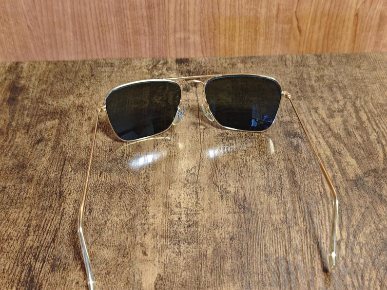 Puede incluir: Gafas de sol con montura dorada y lentes oscuras. Las gafas tienen un dise&ntilde;o cl&aacute;sico de aviador, con doble puente y varillas finas. Las lentes son rectangulares y contrastan con la montura dorada. Est&aacute;n sobre una superficie de madera.