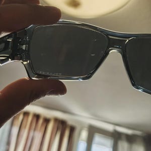 May include: Clear-framed sunglasses with dark lenses. The sunglasses have a rectangular shape and are held up by a hand. The lenses have a slight gradient and the frame is a transparent, light blue color.