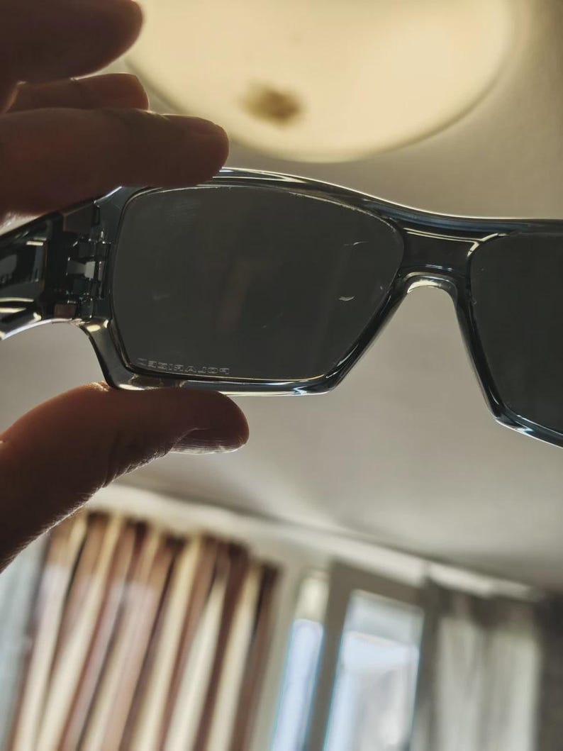 May include: Clear-framed sunglasses with dark lenses. The sunglasses have a rectangular shape and are held up by a hand. The lenses have a slight gradient and the frame is a transparent, light blue color.