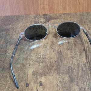 May include: Silver-framed sunglasses with oval lenses. The lenses are dark, and the temples have a mottled gray and black pattern. The sunglasses are resting on a wooden surface.