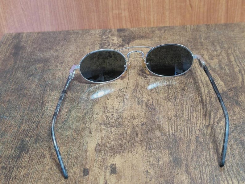 May include: Silver-framed sunglasses with oval lenses. The lenses are dark, and the temples have a mottled gray and black pattern. The sunglasses are resting on a wooden surface.