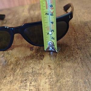 May include: Black sunglasses with dark lenses. The frame measures approximately 2 inches in height. The sunglasses are on a wooden surface.