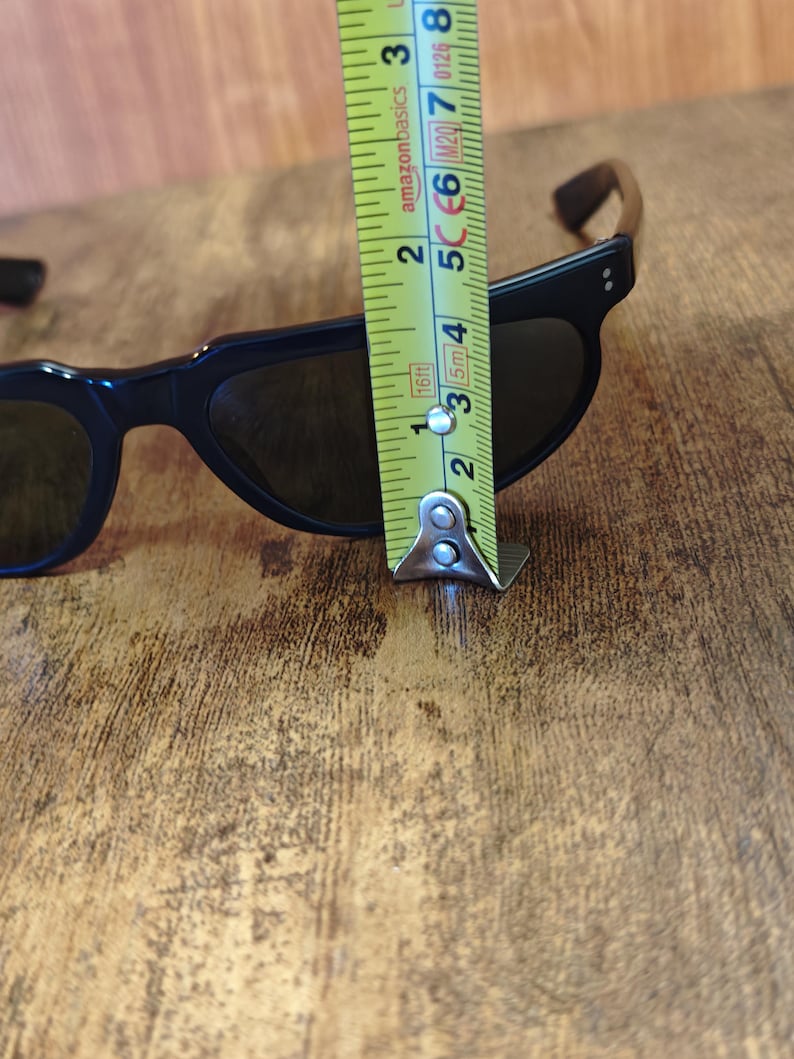 May include: Black sunglasses with dark lenses. The frame measures approximately 2 inches in height. The sunglasses are on a wooden surface.