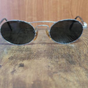 May include: Oval-shaped sunglasses with a silver metal frame and dark lenses. The sunglasses have a double bridge and a classic design. The lenses are dark, providing shade and protection from the sun.