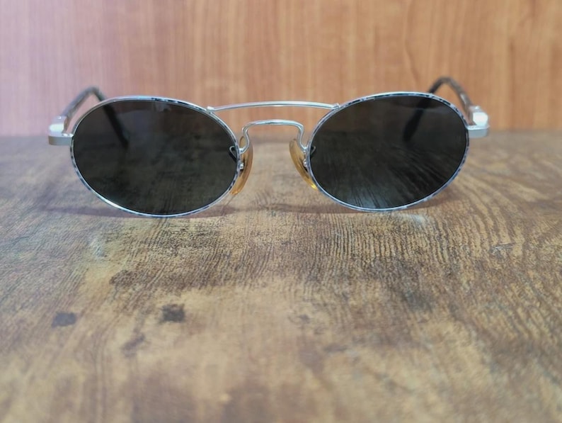 May include: Oval-shaped sunglasses with a silver metal frame and dark lenses. The sunglasses have a double bridge and a classic design. The lenses are dark, providing shade and protection from the sun.