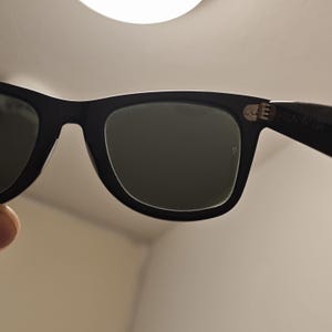 Puede incluir: Gafas de sol negras con lentes tintadas en verde oscuro y una montura de pl&aacute;stico gruesa. La patilla presenta una bisagra de metal plateado visible con un peque&ntilde;o detalle de remache y texto impreso en el interior cerca de la bisagra.