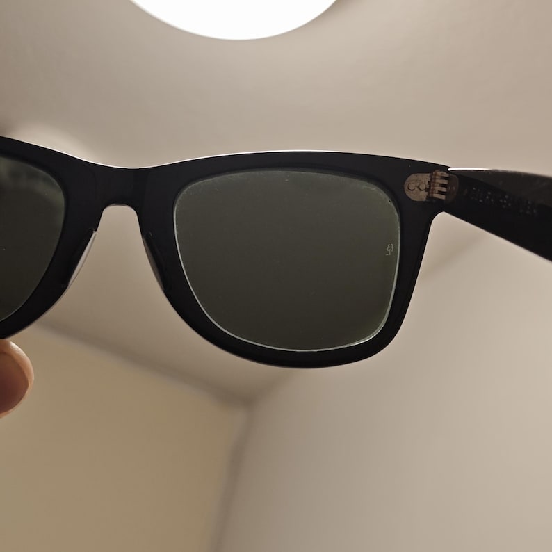 Puede incluir: Gafas de sol negras con lentes tintadas en verde oscuro y una montura de pl&aacute;stico gruesa. La patilla presenta una bisagra de metal plateado visible con un peque&ntilde;o detalle de remache y texto impreso en el interior cerca de la bisagra.