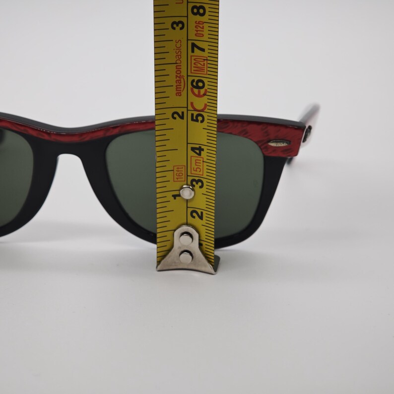 May include: Black sunglasses with a red and black patterned frame. The lenses are green. The sunglasses are being measured with a yellow tape measure. The sunglasses are approximately 5 cm in height.