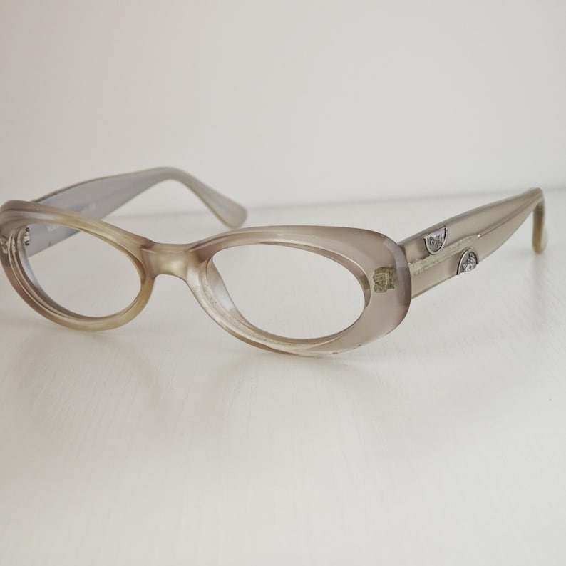 May include: Translucent taupe-colored eyeglasses with an oval frame shape. The temples feature two small, circular silver-toned metal accents embedded on the side. The frames have a smooth, glossy finish and clear lenses, showcasing a simple design.