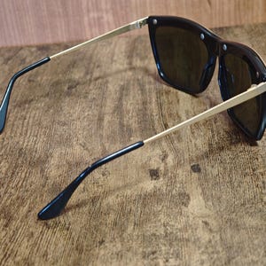 Peut inclure: Lunettes de soleil noires avec des branches dor&eacute;es et des verres fonc&eacute;s. Les lunettes ont une forme carr&eacute;e et sont pos&eacute;es sur un fond en bois. Les branches sont fines et se terminent par des embouts noirs.