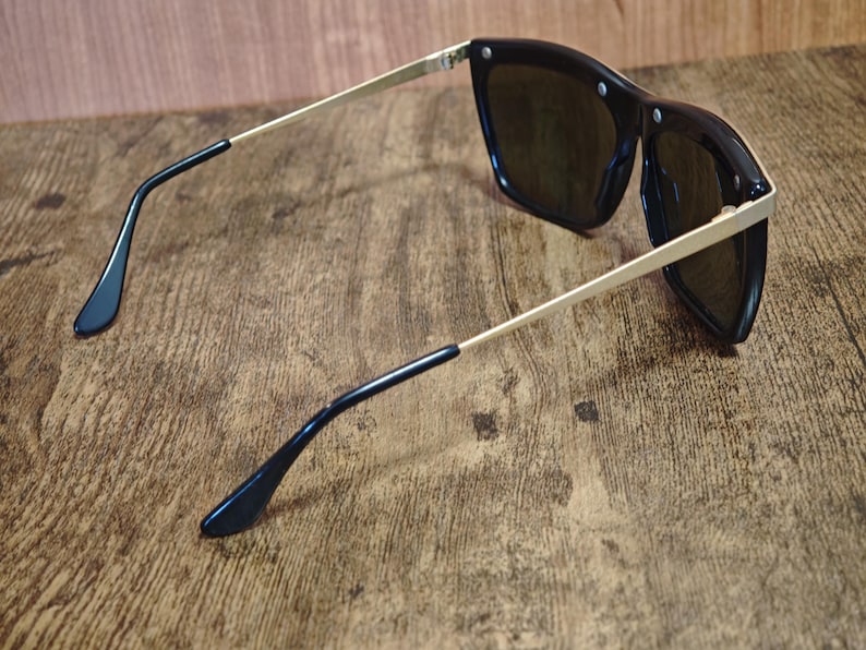 Peut inclure: Lunettes de soleil noires avec des branches dor&eacute;es et des verres fonc&eacute;s. Les lunettes ont une forme carr&eacute;e et sont pos&eacute;es sur un fond en bois. Les branches sont fines et se terminent par des embouts noirs.