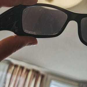 K&ouml;nnte beinhalten: Schwarze Sonnenbrille mit rechteckigem Rahmen und dunklen Gl&auml;sern. Der Markenname "DUCATI" ist wei&szlig; auf dem linken Glas aufgedruckt. Die Sonnenbrille wird von einer Hand gehalten.