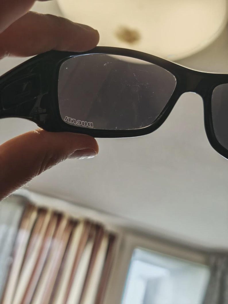 K&ouml;nnte beinhalten: Schwarze Sonnenbrille mit rechteckigem Rahmen und dunklen Gl&auml;sern. Der Markenname "DUCATI" ist wei&szlig; auf dem linken Glas aufgedruckt. Die Sonnenbrille wird von einer Hand gehalten.
