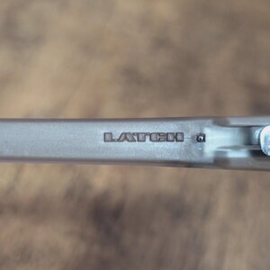 May include: Close-up of a silver-colored metal object with the word "LATCH" embossed on its surface. The object appears to be a component of a larger mechanism, possibly a clasp or fastener. The background is a blurred wooden surface.