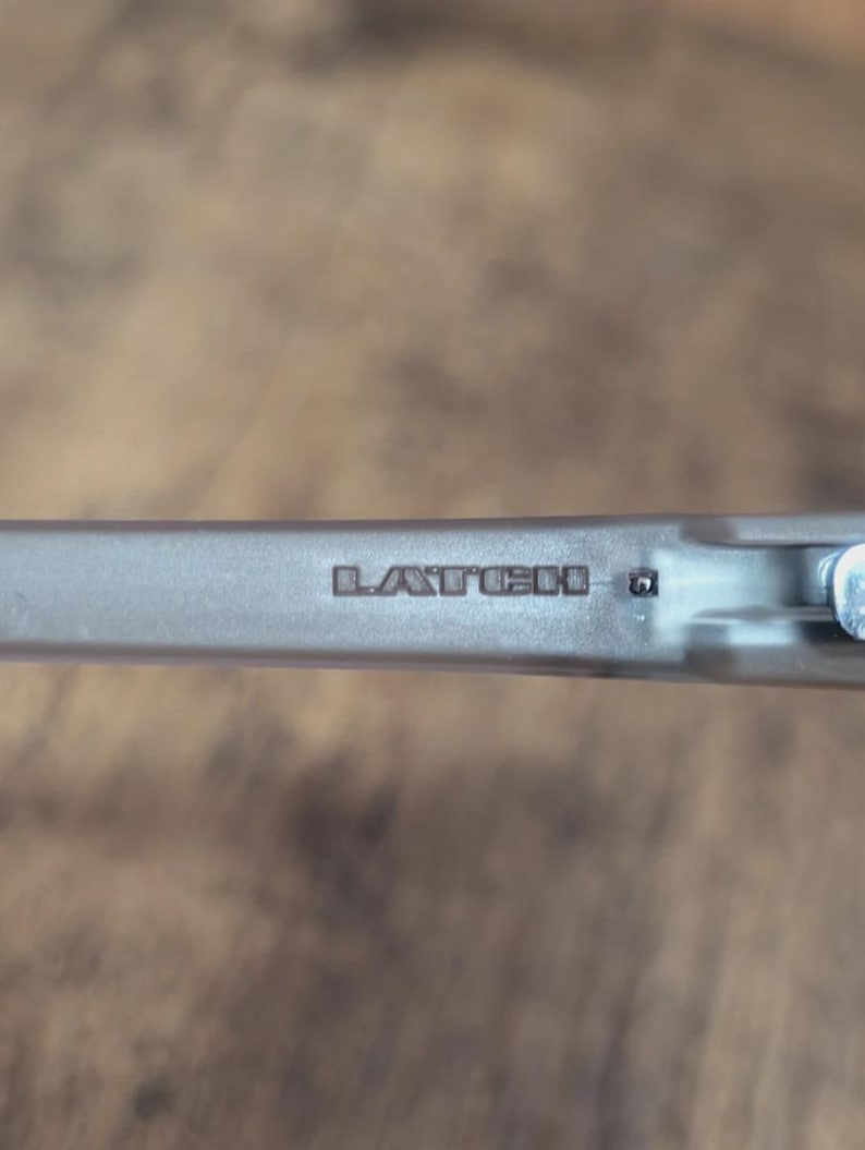 May include: Close-up of a silver-colored metal object with the word "LATCH" embossed on its surface. The object appears to be a component of a larger mechanism, possibly a clasp or fastener. The background is a blurred wooden surface.