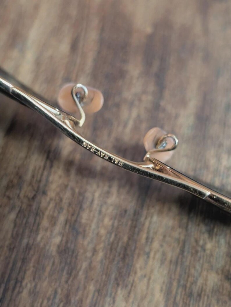 May include: Close-up of a pair of gold-colored eyeglasses. The nose pads are a light peach color. The text "Ray-Ban" is visible on the frame.