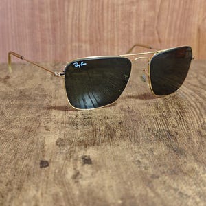 Puede incluir: Gafas de sol con montura dorada y lentes oscuras. El logotipo de Ray-Ban es visible en la lente superior izquierda. Las gafas de sol est&aacute;n sobre un fondo de madera.