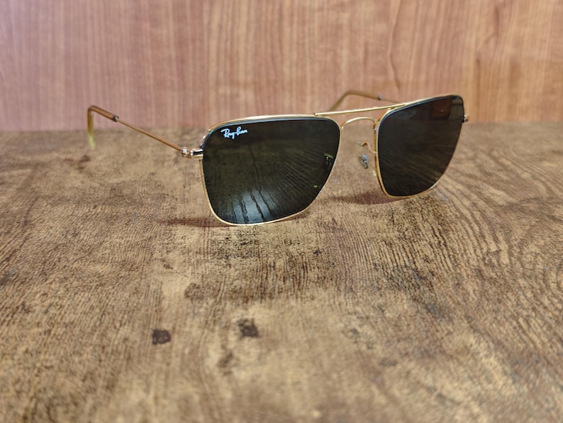 Puede incluir: Gafas de sol con montura dorada y lentes oscuras. El logotipo de Ray-Ban es visible en la lente superior izquierda. Las gafas de sol est&aacute;n sobre un fondo de madera.