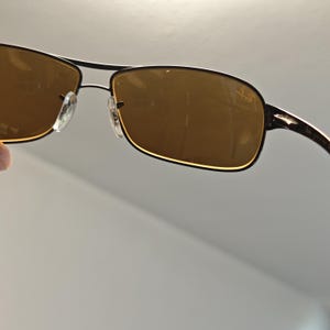 May include: Sunglasses with a dark metal frame and brown tinted lenses. The lenses have a rectangular shape with rounded corners. The bridge of the sunglasses has a double bar design. The temples are thin and curve around the ears.