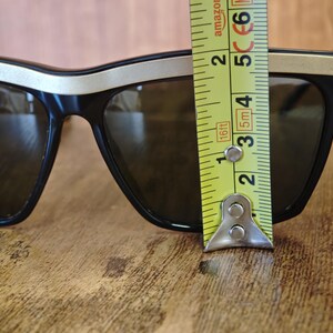 Peut inclure: Lunettes de soleil noires avec une bordure dor&eacute;e sur le dessus et les branches. Les lunettes mesurent environ 5 cm de hauteur. Les verres sont de couleur fonc&eacute;e et r&eacute;fl&eacute;chissante. Sur une surface en bois.