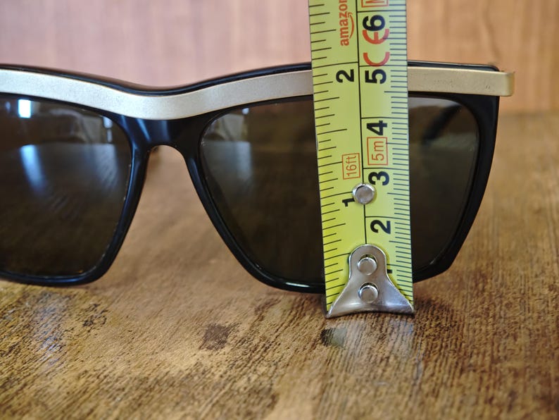 Peut inclure: Lunettes de soleil noires avec une bordure dor&eacute;e sur le dessus et les branches. Les lunettes mesurent environ 5 cm de hauteur. Les verres sont de couleur fonc&eacute;e et r&eacute;fl&eacute;chissante. Sur une surface en bois.