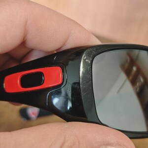 K&ouml;nnte beinhalten: Nahaufnahme einer schwarzen Sonnenbrille mit rotem Akzent am Rahmen. Die Gl&auml;ser sind reflektierend und der Rahmen weist Gebrauchsspuren auf. Die Sonnenbrille wird von einer Hand gehalten.