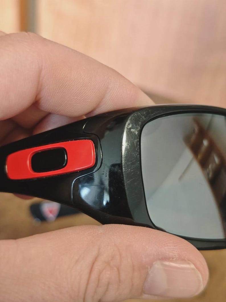 K&ouml;nnte beinhalten: Nahaufnahme einer schwarzen Sonnenbrille mit rotem Akzent am Rahmen. Die Gl&auml;ser sind reflektierend und der Rahmen weist Gebrauchsspuren auf. Die Sonnenbrille wird von einer Hand gehalten.